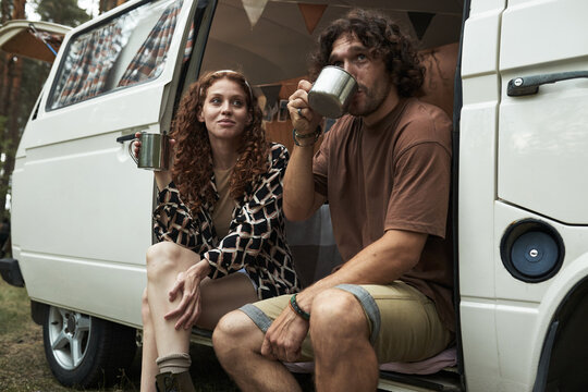 Couple sitting at a vintage van in forest campsite environment, enjoying coffee and relaxing in rural setting. Both individuals are smiling and seem comfortable in natural surroundings