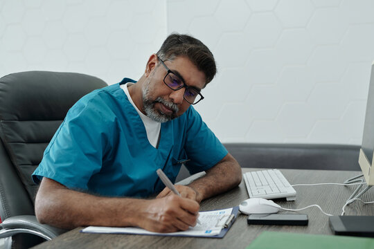 Mature Biracial Doctor Sitting at Desk Filling Medical Record Form