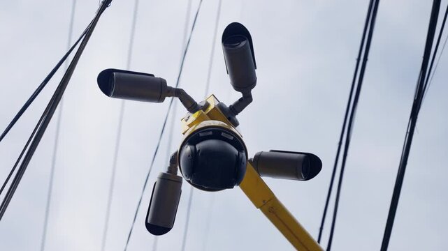 Close-up view of a multi-camera surveillance system mounted on a yellow pole amid overhead utility wires, designed for comprehensive public monitoring and security.