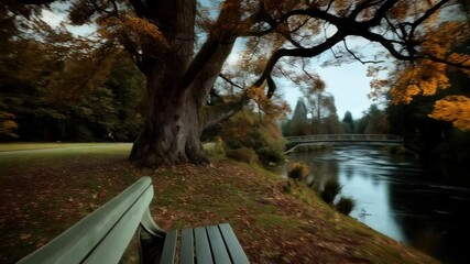 Autumn scene depicts a park bench overlooking river flowing under small footbridge surrounded by trees with golden fall foliage.