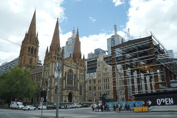 Naklejka premium A wide shot captures a historic cathedral with two prominent spires standing beside a modern building under construction, with a city skyline visible in the background under a cloudy sky. Cars and ped