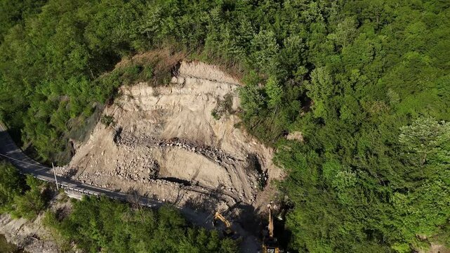 Drone shot of SP21 Vernasca after rockslide on April 22nd 2025, excavators operate to clear blocked road, surrounded by woodland and scattered rocks, real time