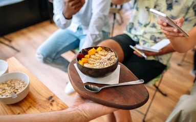 Unrecognizable chef showing vegan breakfast bowl made with mango, banana, raspberries, cereals and muesli to food bloggers and influencers taking pictures with smartphones in cooking master class.