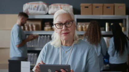 Tilt up portrait of elderly female volunteer in blue T-shirt using digital tablet and looking at camera at donation center - Powered by Adobe
