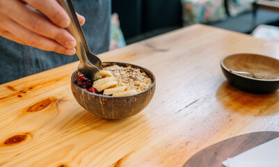 Close up of unrecognizable chef hands preparing vegetarian breakfast putting banana slices on coconut bowl using metal tongs. Concept of healthy vegan dish made with raspberries, cereals and muesli.