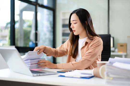 Asian businesswoman focused on sorting stacks of paperwork at her desk with a laptop and coffee cup. - Powered by Adobe