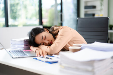 verworked Asian woman falls asleep on desk with paperwork, laptop, and coffee. Office fatigue, deadline stress, and burnout concept