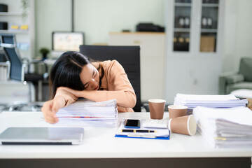 verworked Asian woman falls asleep on desk with paperwork, laptop, and coffee. Office fatigue, deadline stress, and burnout concept