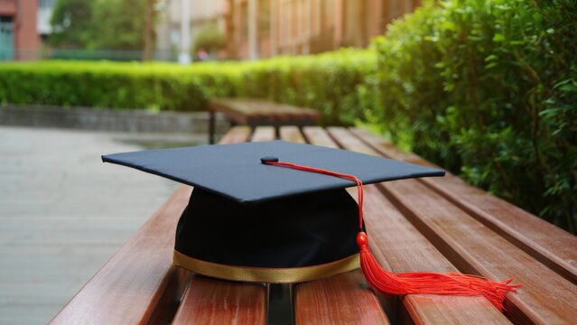 Graduation mortarboard with red tassel placed on wooden bench in campus courtyard, associated with commencement ceremony, academic achievement, higher education, and student milestone events