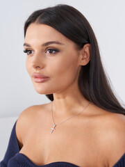 Brunette woman with long hair gazes to the side, wearing cross necklace and off-shoulder top....