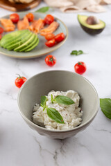 Top view of ricotta cheese in gray bowl with basil leaves, on light background with blurred cherry tomatoes and avocado. Clean and natural style. Concept of healthy cooking and food minimalism