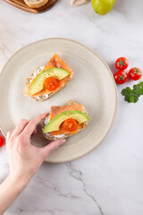 Top view of two healthy toasts with avocado, salmon and cherry tomatoes on a beige plate. A female hand holds one of the toasts. Concept of healthy breakfast and modern lifestyle