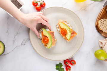 Female hand holding a slice of healthy toast with avocado, smoked salmon and cherry tomato, served on a ceramic plate. Light marble background with fresh ingredients. Concept of healthy breakfast 