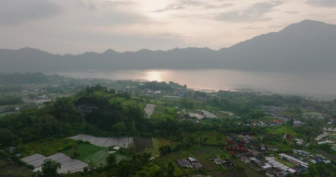 4K drone shot at sunrise over Lake Danau Batur in Kintamani, with Gunung Abang in the distance, traditional lake farming along the shore, and golden sun flare in frame