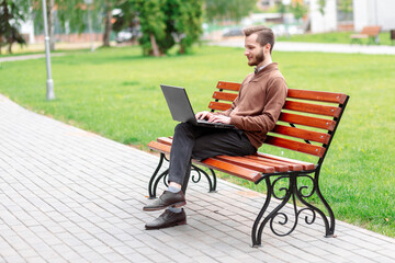 Happy young man sitting on bench in green park and working on laptop