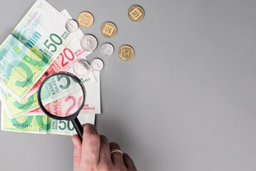 Israel currency 100 and 50 New Israeli shekel (NIS) banknotes and agorot coins under magnifying glass, grey background, concept of inflation, economic crisis, rising prices, and financial management.