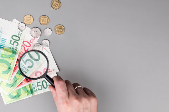 Israel currency 100 and 50 New Israeli shekel (NIS) banknotes and agorot coins under magnifying glass, grey background, concept of inflation, economic crisis, rising prices, and financial management.