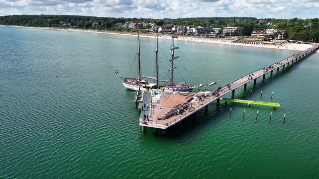 Aerial footage of sailing ship docked at the long pier in Scharbeutz on the Baltic Sea with pan and retreat movement of the camera. Wooden pier with three-masted sailing vessel at seaside resort.