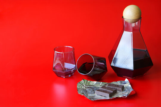 Tinted glass decanter with matching tumblers and foil-wrapped chocolate pieces arranged on a bold red backdrop – ideal for alcohol branding, gourmet packaging, or party menu visuals. Photo - Powered by Adobe
