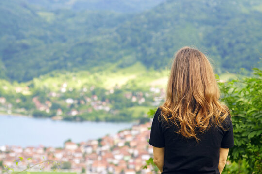 Photographed from behind, a woman with wavy golden-red hair enjoys the view of red-tiled houses built by the water and mountains. A female tourist at a viewpoint in Zvornik, Bosnia and Herzegovina.
