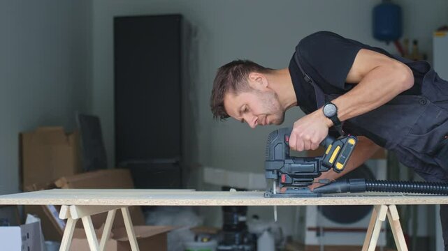 Man in workwear using jigsaw to cut chipboard panel in modern home workshop. Concept of DIY renovation, craftsmanship and home improvement process.