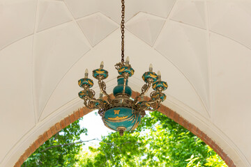 Elegant chandelier suspended from a vaulted ceiling in Tehran, with lush greenery visible in the background, blending architectural beauty with nature.