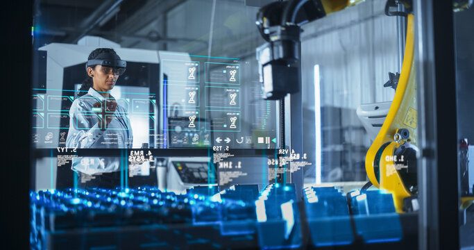 Engineer Wearing Augmented Reality Headset Operates Robotic Arm in a Factory, Interacting With Virtual Controls on a Transparent Holographic Interface. Advanced Machinery and Digital Displays.