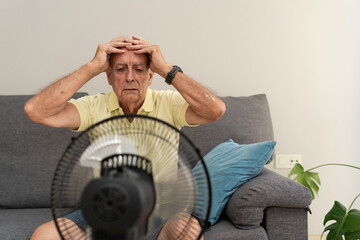 Elderly man with hands on head trying to cool off with electric fan during summer heatwave at home