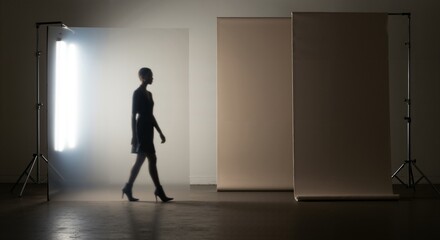 Fototapeta premium Silhouette of an elegant woman walking in a dark photo studio with bright backlight. Fashion model on a professional photoshoot set with backdrops.