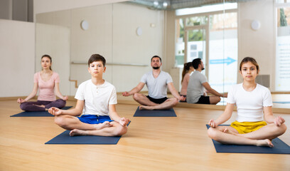 Friendly sporty family of young parents and teenage kids doing yoga together in fitness studio, sitting on mats in lotus position and meditating