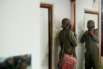 Man standing in front of mirror wearing uniform with backpack and contemplating his reflection. Indoor setting with minimalistic decor highlighting the scene