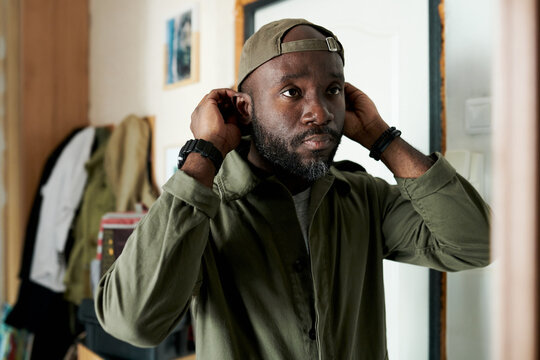Black person adjusting cap, standing in room with shelves and pictures in background. Focused and intent expression, casual indoor setting