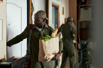 Man carrying groceries entering front door with reflection visible in mirror. Holding a paper bag filled with fresh produce and bread, fully focused on opening the door