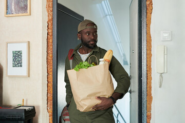 Man entering modern home while carrying grocery bag filled with vegetables and baguette, emphasizing healthy lifestyle and daily routine