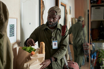 Man holding grocery bag filled with fresh produce and a baguette, wearing uniform and name badge, with mirror reflecting part of room in background