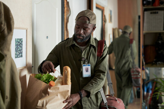 Man holding grocery bag filled with fresh produce and a baguette, wearing uniform and name badge, with mirror reflecting part of room in background