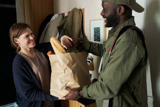 Smiling woman accepting a paper bag filled with fresh groceries from a cheerful delivery person standing by doorway, creating positive interaction between client and service provider