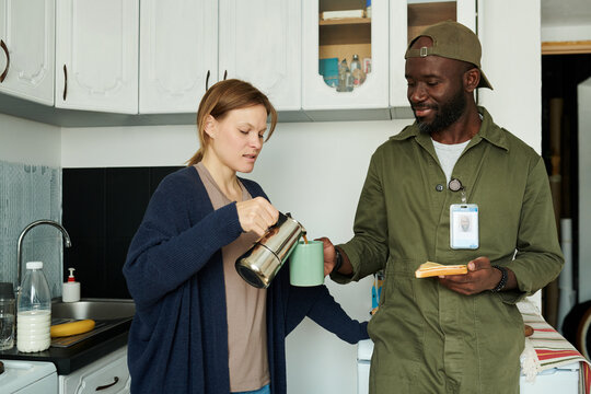 Biracial couple spending time in kitchen with woman pouring coffee and man holding toast, showing friendly interaction. Modern kitchen furnishings in background - Powered by Adobe