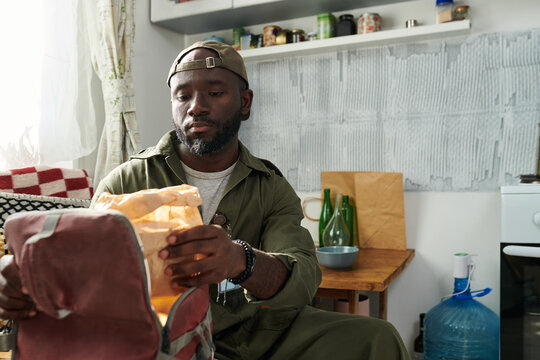 Man wearing a cap is seen packing lunch into a brown bag in a kitchen setting, shelves filled with various items visible in the background
