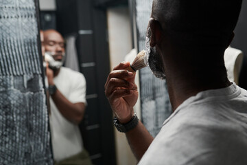 Man shaving beard while standing in front of bathroom mirror. Reflection showing details of grooming routine in a modern bathroom setting
