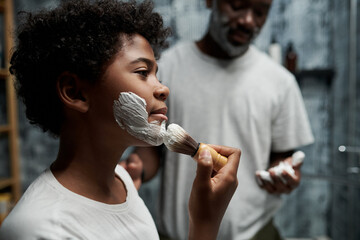 Father teaching his young son proper shaving techniques in bathroom. Son focusing and learning while father provides guidance and support with smile