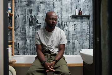 Black man sitting in modern bathroom with textured wallpaper, looking deep in thought and reflecting in space filled with contemporary design elements