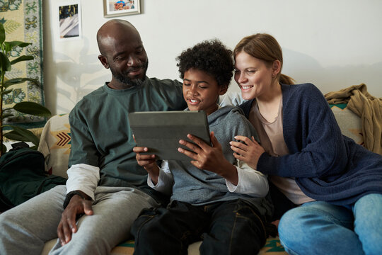 African American father, Caucasian mother, and their son smiling and sitting on a couch looking at a tablet screen, enjoying family time