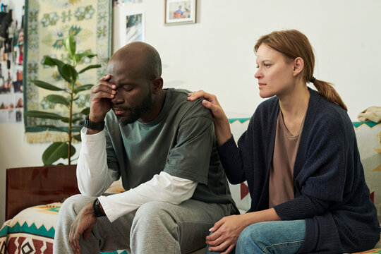 Woman sitting beside man on couch, offering comfort in supportive, emotional moment, with cozy living room decor and framed photos visible in background