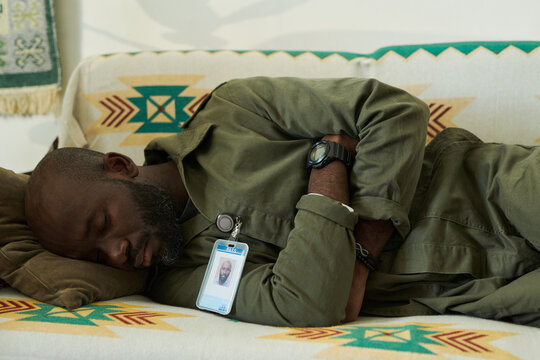 Man in olive green jacket peacefully sleeping on couch with patterned blanket in background while wearing an ID badge and watch on wrist