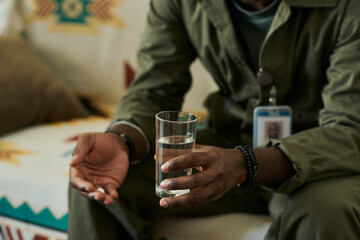 Close-up of mans hands holding glass of water and pills while sitting on couch in cozy living space. Man is wearing casual clothing with ID badge on neck