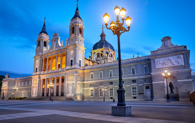 Madrid, Spain. Cathedral Santa Maria la Real de la Almudena at Plaza de la Armeria. Famous landmark with sunset sun, flowers and green bush. Street lamps with illumination and picturesque blue sky.