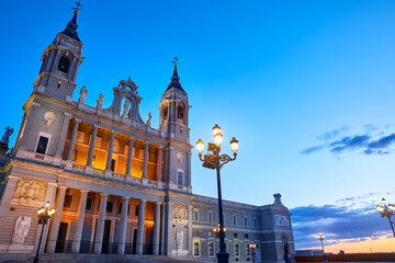 Madrid, Spain. Cathedral Santa Maria la Real de la Almudena at Plaza de la Armeria. Famous landmark with sunset sun, flowers and green bush. Street lamps with illumination. Low angle view.