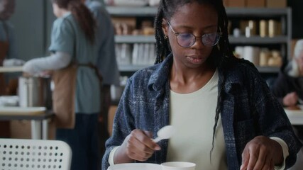 Rack focus of young African American woman eating hot meal while sitting at table at food donation center with volunteers