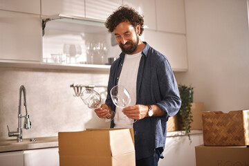 A man is unpacking moving boxes in a bright kitchen, smiling as he holds two wine glasses. Sunlight...